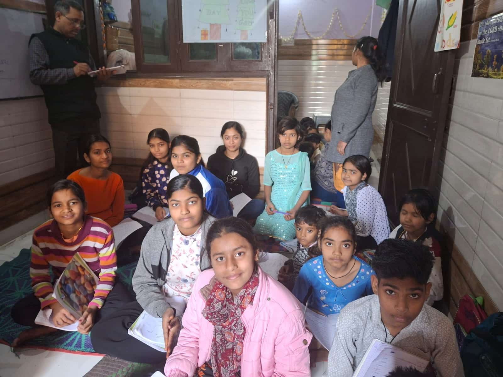 A group of students is sitting closely together on the floor in a classroom, writing in notebooks during a study session