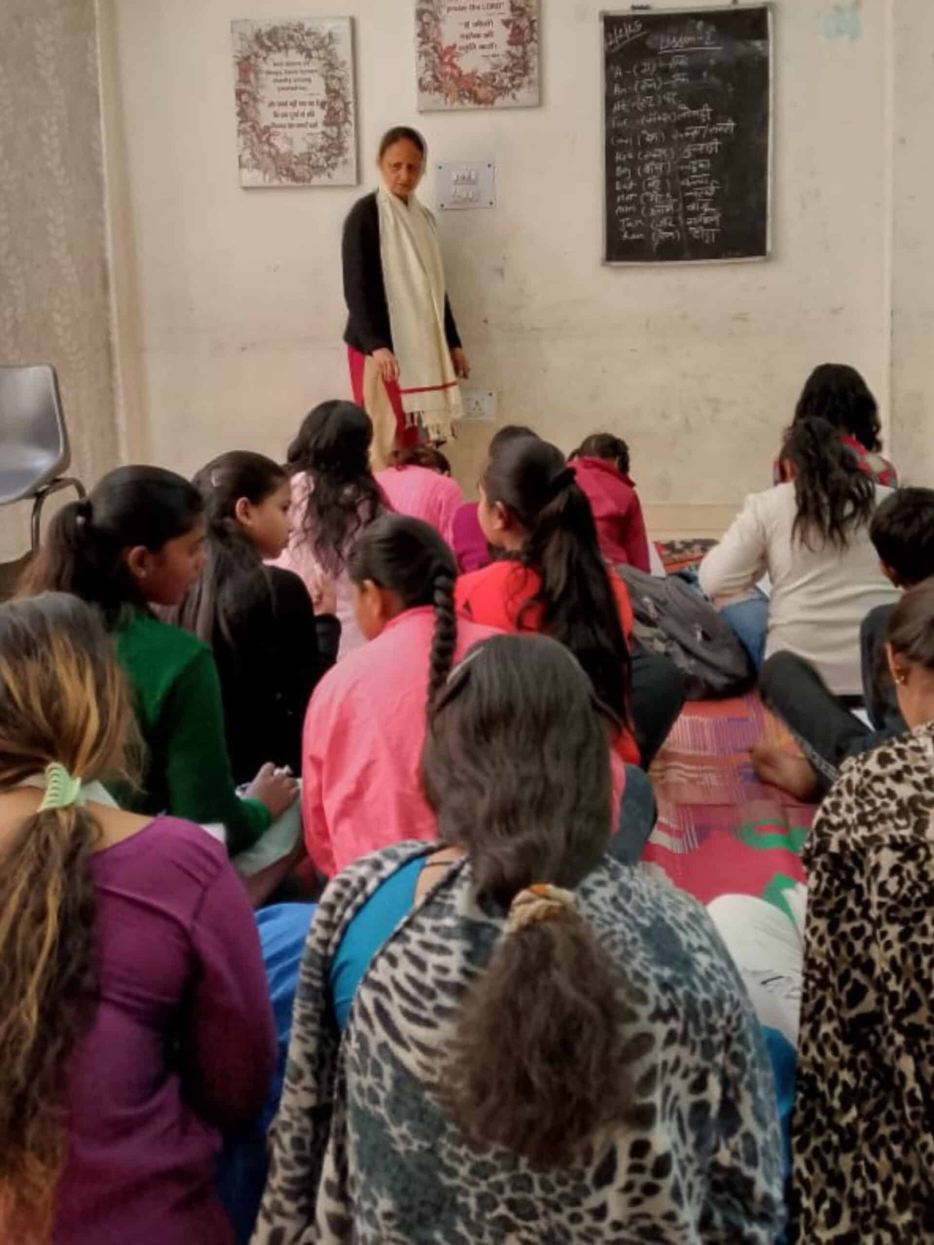 A teacher stands at the front of a classroom near a blackboard, instructing a group of girls seated on the floor as they write in their notebooks during a lesson.