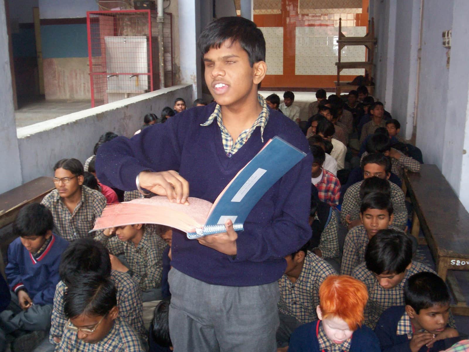 A student stands in front of a classroom holding open a notebook, while classmates sit on the floor behind him.