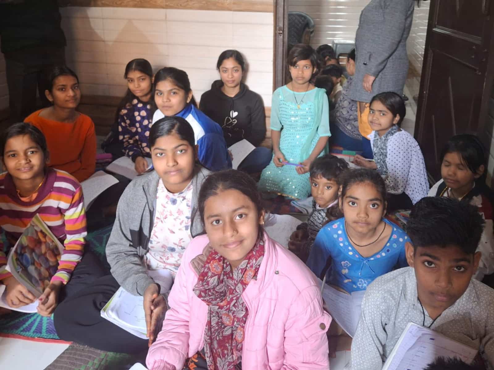 A group of students is sitting closely together on the floor in a classroom, writing in notebooks during a study session