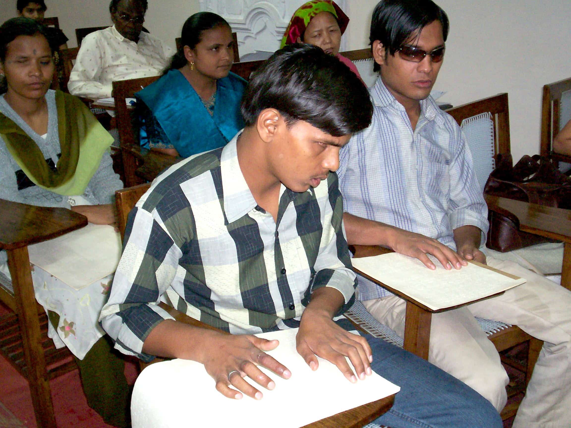 A group of students sits in a classroom, reading braille on sheets placed on their desks.