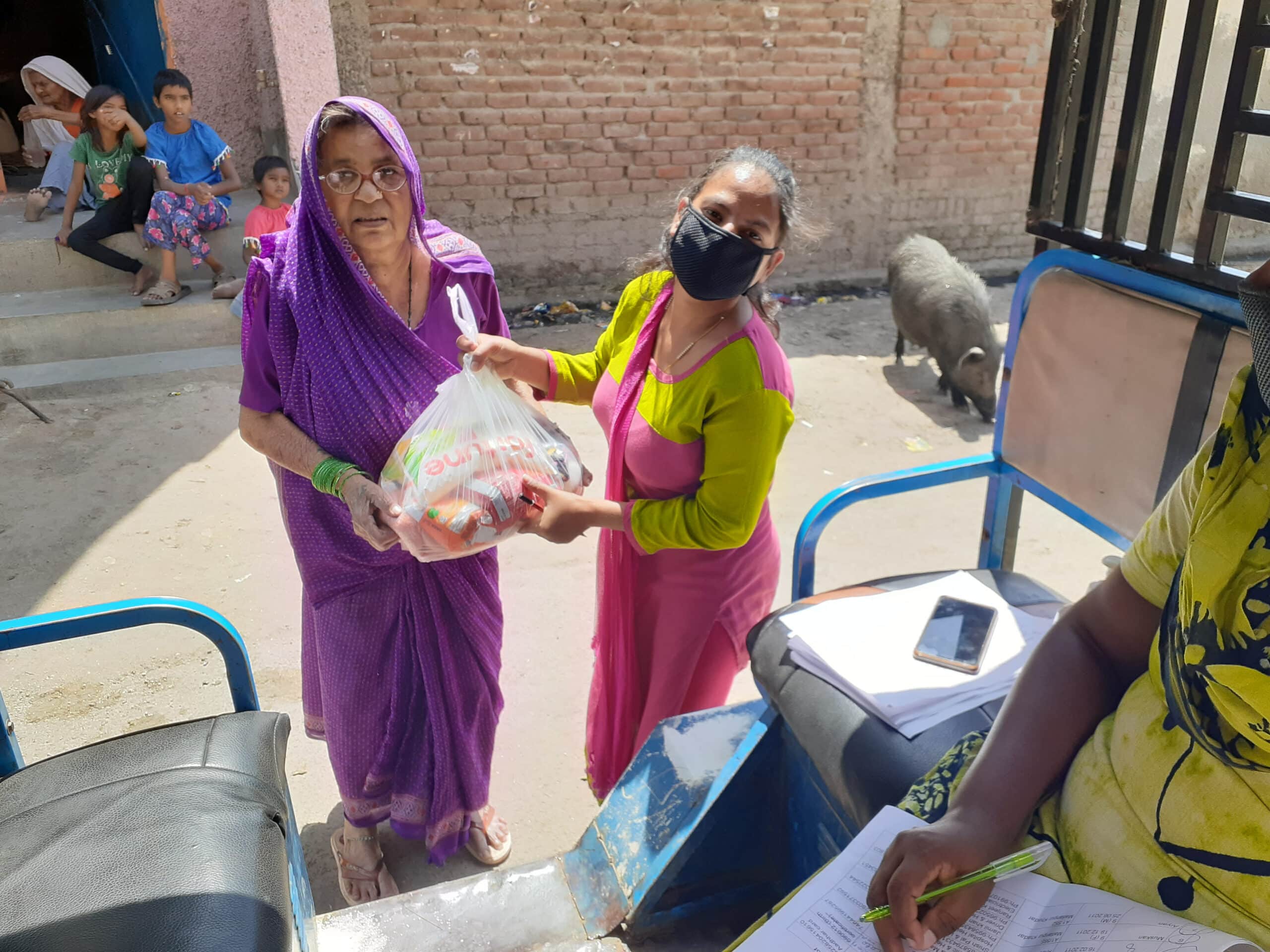 Two women exchange a grocery bag outdoors while a person records details and children sit in the background.