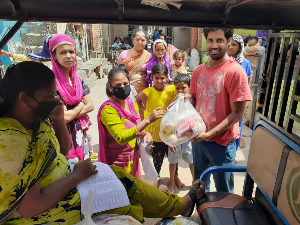 A woman in a yellow saree takes notes on a clipboard while another woman hands a grocery bag to a smiling man in a street setting, surrounded by children and community members.