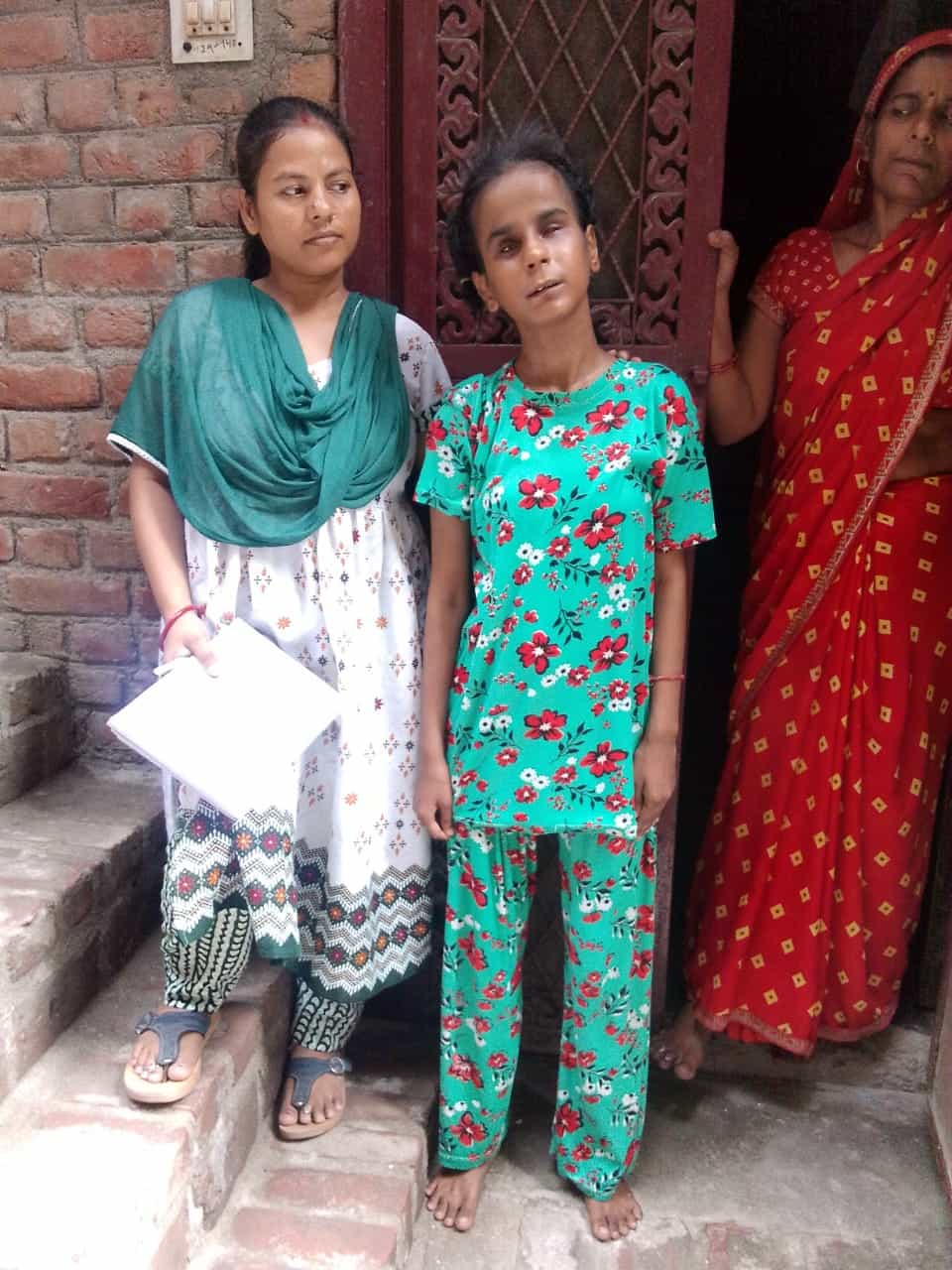 Three women stand outside a brick building. The central woman appears blind, and another holds papers, while the third looks on.