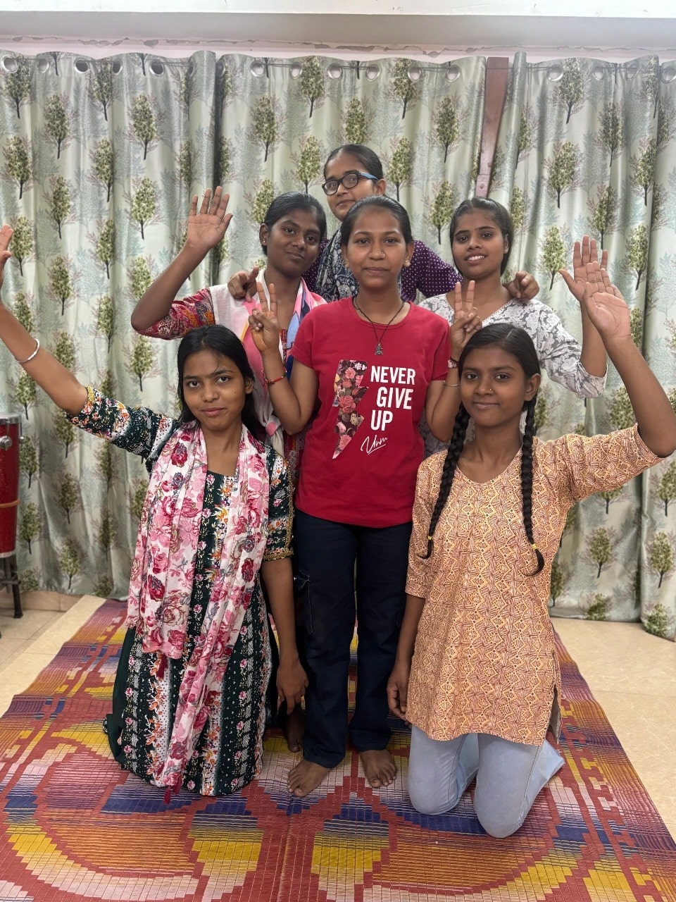 Six girls indoors, some standing and some kneeling on a colorful floor mat, raising their hands in playful gestures. One girl in the center wears a red T-shirt with the words 'Never Give Up.' The background features patterned curtains with a tree motif.