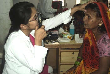 Healthcare worker in a white coat conducts a medical examination of a woman wearing a colorful sari, inside a clinic room with a desk, medical supplies, and paperwork in the background.