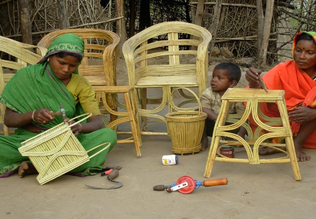 Women and children sitting outdoors weaving and constructing furniture with bamboo or cane, surrounded by finished chairs and tools, as part of a rural livelihood restoration program.