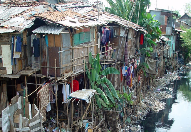Row of makeshift houses constructed from tin, wood, and scrap materials, built on stilts along a polluted riverbank with visible garbage, in a densely populated urban area.